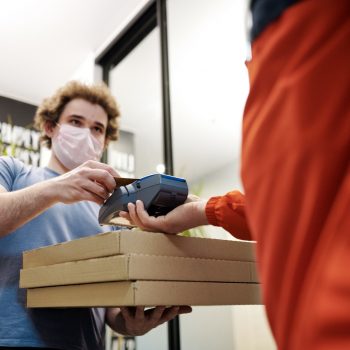 A man wearing a face mask holding pizza boxes using a card reader machine held by another person